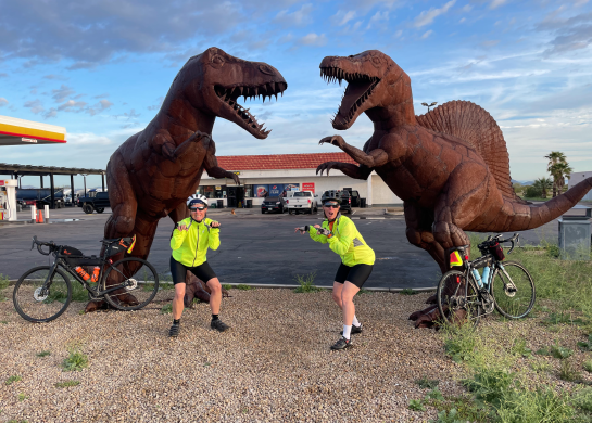 Photo of two women in biking clothes posing in front of two dinosaur statues. 