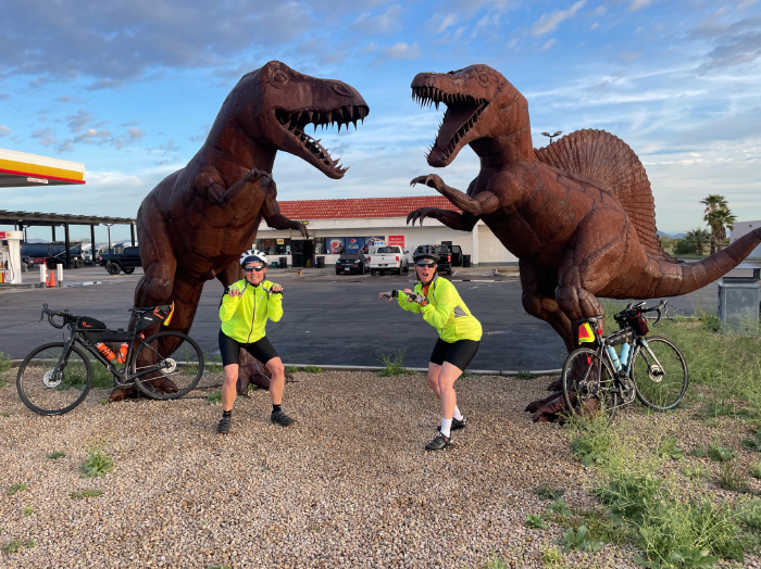 Photo of two women in biking clothes posing in front of two dinosaur statues. 
