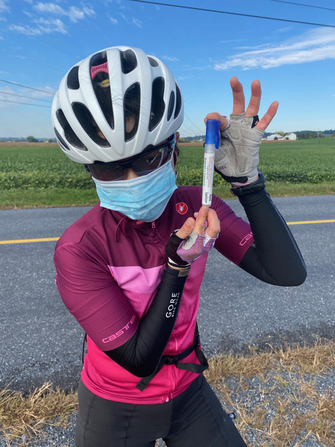 A woman in bicycling gear is wearing a surgical mask while standing by the side of the road.