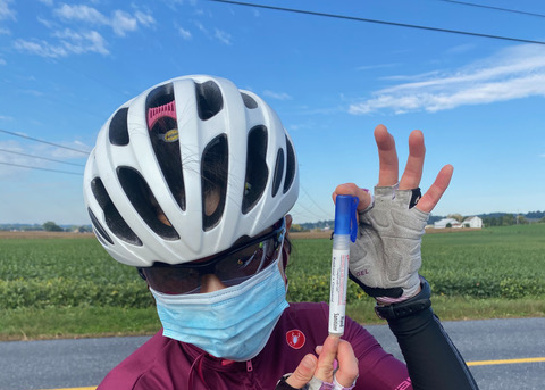 A woman in bicycling gear is wearing a surgical mask while standing by the side of the road.