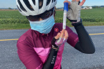 A woman in bicycling gear is wearing a surgical mask while standing by the side of the road.